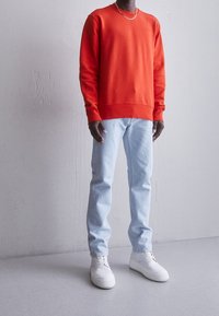 Červená mikina, světlé modré džíny a bílé tenisky. Mikina má kulatý výstřih a žebrované manžety; džíny mají rovný střih.