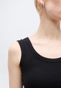 Κοντινό πλάνο ενός ατόμου που φοράει μαύρο ριπ μπλουζάκι χωρίς μανίκια και χρυσό κρίκο σκουλαρίκι, με λαιμό και ώμο να φαίνονται μπροστά σε ανοιχτό φόντο.