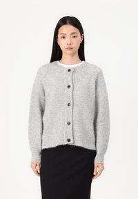 Cardigan texturé gris clair avec un col rond, doté de sept boutons foncés sur le devant et de poignets côtelés. Porté par-dessus une jupe noire.