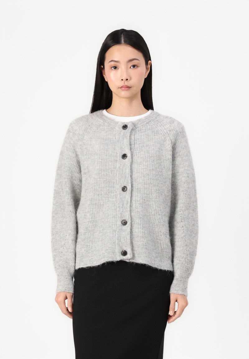 Cardigan texturé gris clair avec un col rond, doté de sept boutons foncés sur le devant et de poignets côtelés. Porté par-dessus une jupe noire.
