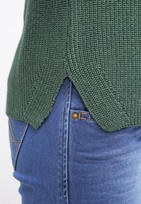 Pull en maille vert foncé avec fente latérale porté par-dessus un jean en denim bleu, montrant les détails de la poche et des coutures.