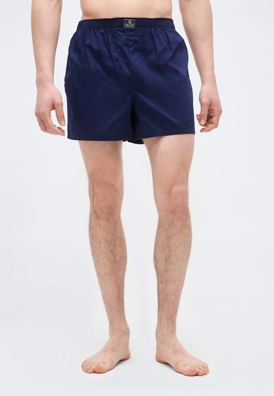 Άνδρας μοντέλο που φοράει βαμβακερά μπόξερ σορτς με ελαστική μέση σε ναυτικό μπλε, όρθιος ξυπόλυτος μπροστά σε λευκό απλό φόντο.