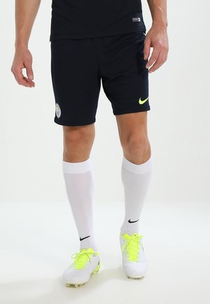 Mand iført sorte sportsshorts med Manchester City- og neon Nike-logoer, hvide knæstrømper og hvide fodboldestøvler med neon-snørebånd.