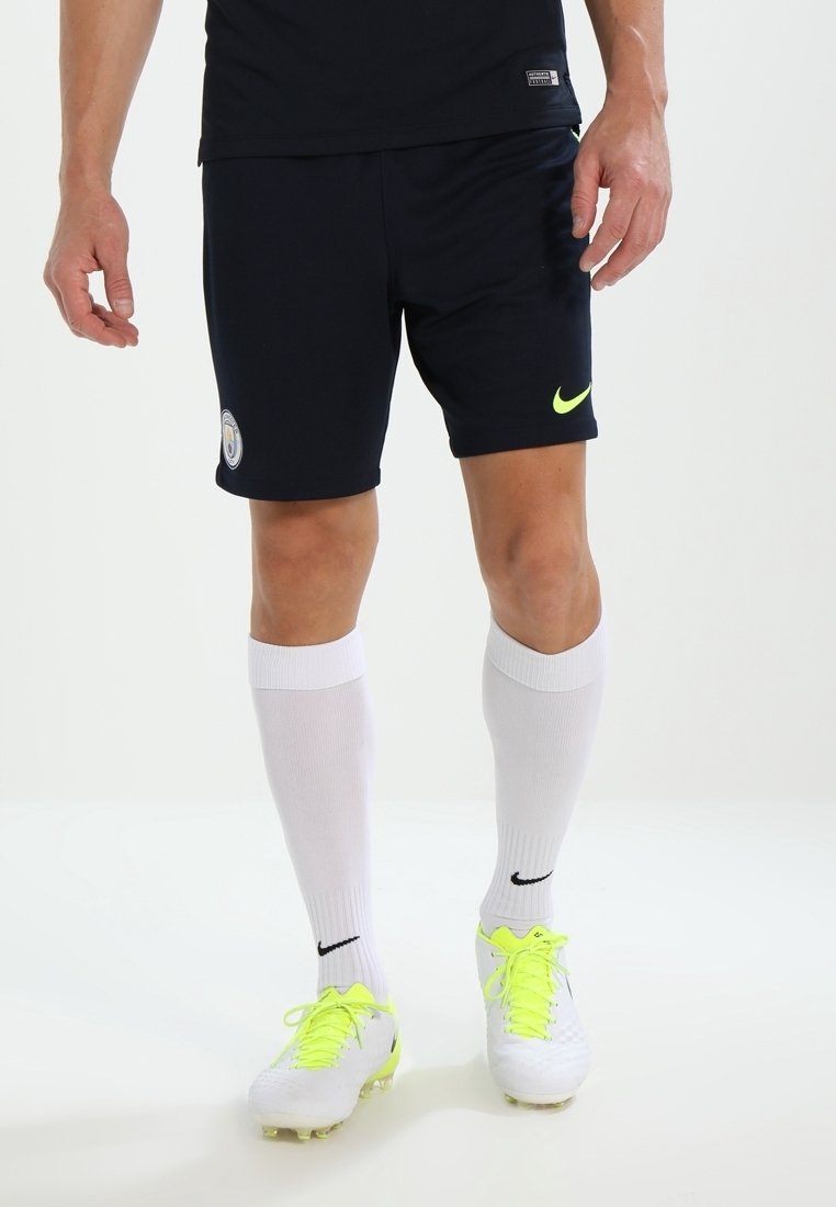Homme portant un short de sport noir avec les logos de Manchester City et Nike en néon, des chaussettes blanches montantes et des chaussures à crampons blanches avec des lacets néon.