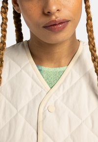 Gilet en crème matelassé avec un motif en losange et des finitions beiges, présentant un col rond et une couche texturée verte en dessous.