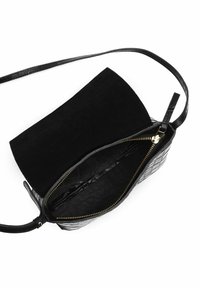 Czarna skórzana torebka na ramię z wzorem krokodyla, złoty zamek częściowo otwarty, cienki regulowany pasek na tle białym.