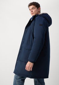 Abrigo acolchado azul marino con capucha, que cuenta con un cierre frontal, botones a presión y dos grandes bolsillos frontales. Textura suave, estilo casual.