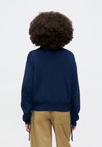 Suéter de malha azul marinho com textura canelada, corte descontraído e laços laterais. Combinado com calças em tom bege, visto de costas.