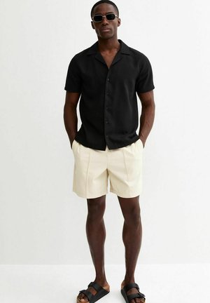 Mężczyzna ubrany w czarną koszulę z krótkim rękawem na guziki, beżowe szorty, czarne sandały i ciemne okulary przeciwsłoneczne, stoi z rękami w kieszeniach.