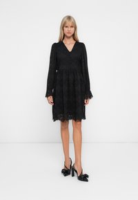 Blonde Frau steht in einem knie-langen schwarzen Spitzenkleid mit langen Ärmeln und V-Ausschnitt, trägt schwarze High Heels mit Schleifendetail.