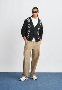 Homme portant un cardigan noir avec des motifs de serpents blancs, un t-shirt blanc, un pantalon large beige, des baskets blanches et des lunettes de soleil noires, se tenant sur un sol bleu.