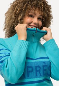 Ljusblå, texturerad sweatshirt med hög krage, dragkedja och mörkblå ränder med ordet "APRÈS" över bröstet.