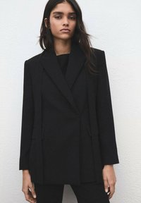 Czarna, dopasowana marynarka o strukturalnym kroju, z jednorzędowym zapięciem, klapami z wycięciami i przednimi kieszeniami. Gładka tekstura z mieszanki wełny.
