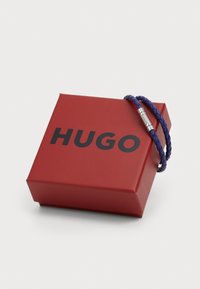 Bracciale in pelle intrecciata viola con chiusura in metallo argentato, poggiato su una scatola rossa con la parola "HUGO" in grassetto lettere nere.
