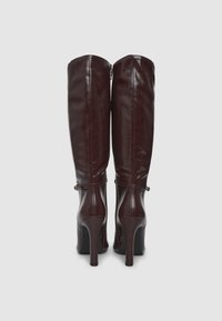 Bottes à genoux bordeaux en simili cuir brillant avec un talon fin. Elles présentent une fermeture éclair sur le côté et un détail de sangle décoratif près de la cheville.