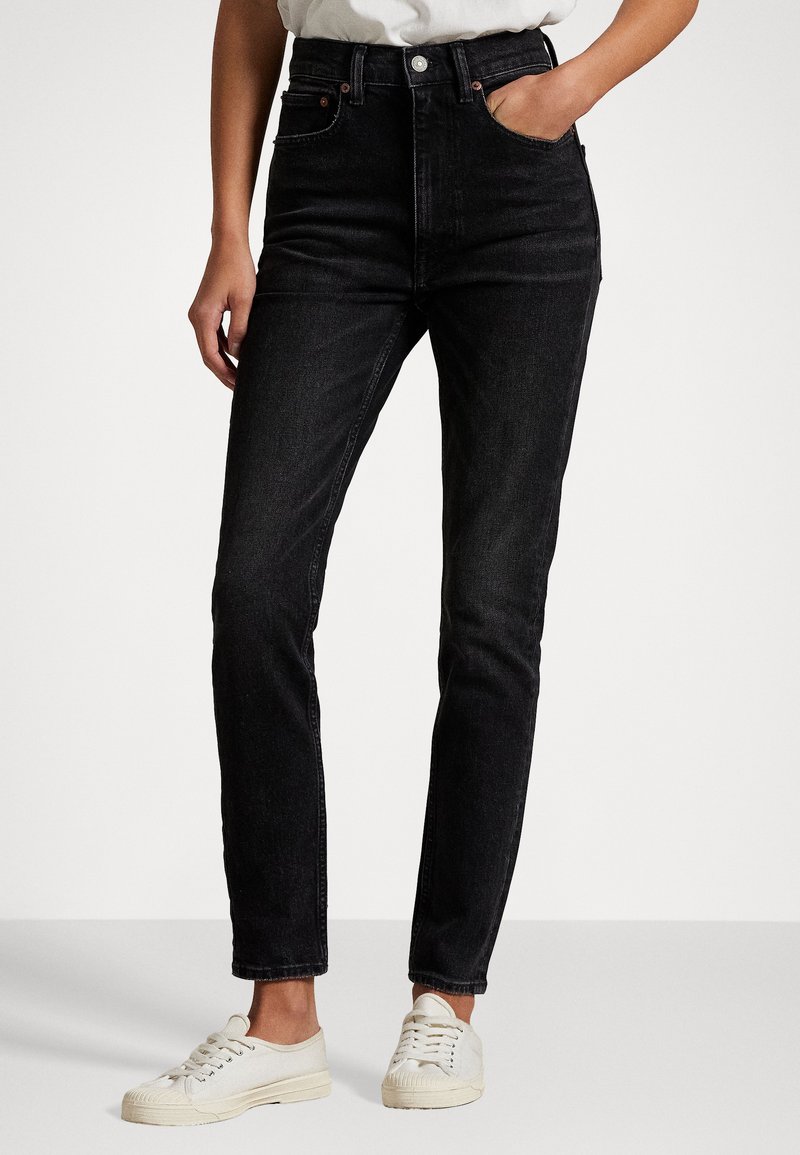 Mujer vestida con jeans negros de tiro alto con bolsillos delanteros, combinados con zapatillas blancas y una camisa clara metida por dentro, de pie sobre un suelo liso.