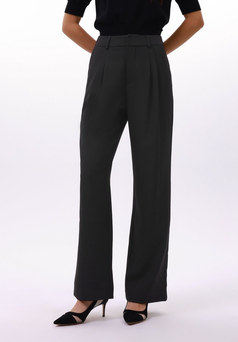 Femme portant un pantalon noir évasé taille haute associé à des escarpins noirs à bout pointu, debout avec les mains dans le dos.