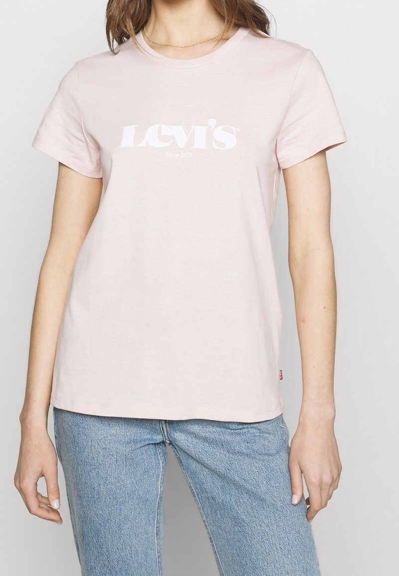 Femme portant un T-shirt Levi's rose pâle avec un logo blanc et un jean bleu clair, debout les bras détendus le long du corps.