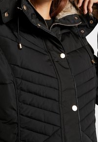 Giubbotto imbottito nero con cappuccio foderato in pelliccia sintetica, cuciture orizzontali e a zig-zag, dettagli in metallo dorato e cordini regolabili.