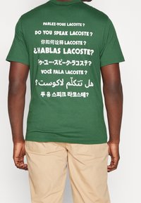 Zelené bavlněné tričko s krátkým rukávem a kulatým výstřihem, na zadní straně s potiskem různých frází v bílé barvě v několika jazycích.