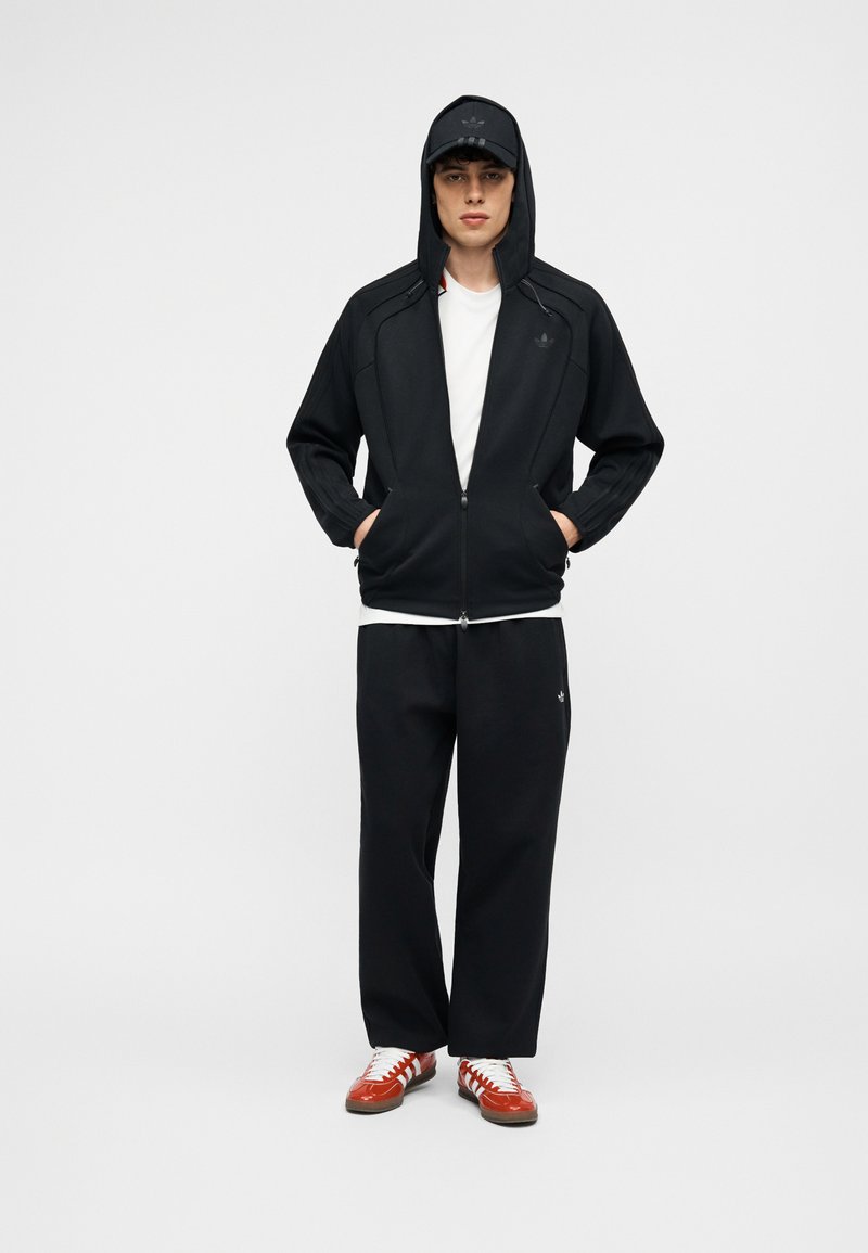 Jonge man die een zwarte hoodie met rits draagt, zwarte broek, wit T-shirt, zwarte pet en rood-witte sneakers, staande met handen in de zakken.