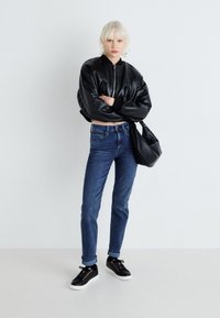 Black leather bomber jacket, blue slim-fit jeans, black sneakers with white soles. Model poses against a neutral background.