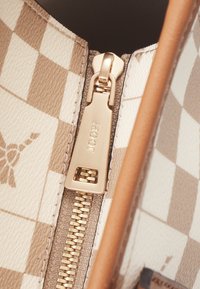 Detailní záběr na béžovou a bílou kostkovanou tašku se zlatým zipem s vyrytým nápisem "JOOP!" a viditelnými detaily šití.