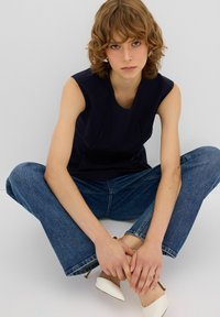 Top senza maniche blu navy con scollatura a forma curva, abbinato a jeans blu. La modella indossa décolleté bianchi a punta, seduta con le gambe incrociate.