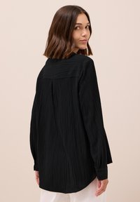 Camicia nera in tessuto crinkle con orlo arrotondato, maniche lunghe, colletto e polsini con bottone. Il retro presenta dettagli testurizzati verticali.