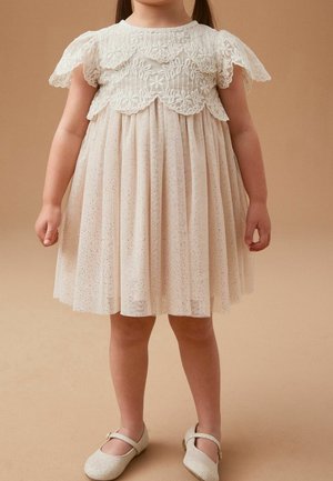 Robe crème clair avec corsage à détails en dentelle, manches courtes à volants et jupe plissée avec des accents pailletés. Portée avec des chaussures beiges scintillantes.