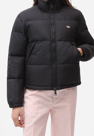 Personne portant une doudoune matelassée noire avec une fermeture éclair frontale et des poches latérales, associée à un pantalon rose clair.