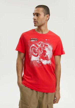 Mężczyzna w czerwonym t-shircie z białą grafiką motocrossowca na motocyklu terenowym oraz napisem "SUPER ENDURO" na klatce piersiowej.