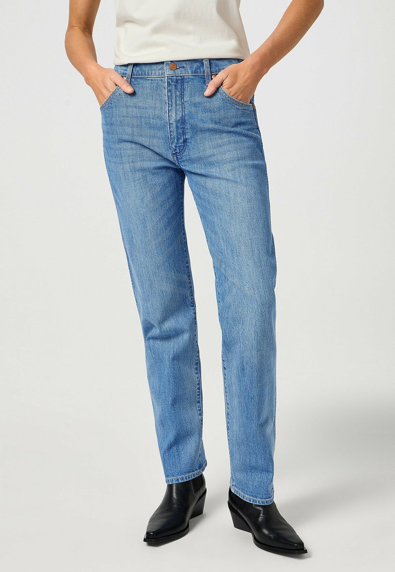 Jeans de denim azul claro con un corte recto, costuras visibles, bolsillos frontales y un cierre clásico de botón. Llevados con botas de tobillo puntiagudas negras.