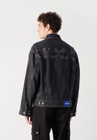 Sort denimjakke med en let struktureret overflade, prydet med et stort prægede logo på ryggen og et blåt mærke i kanten.