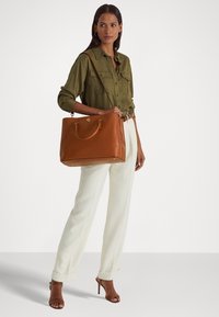 Brown leather tote bag with a structured shape, dual handles, and a gold logo. Paired with an olive green shirt and cream wide-leg pants.