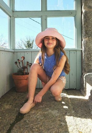 Sombrero rosa claro, top de punto azul, pantalones cortos beige; niño agachado, ajustando zapatos marrón claro en un entorno interior iluminado por el sol.