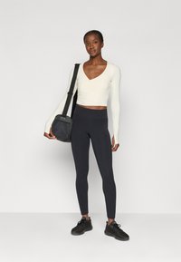 Langemouwige, v-hals crop top in crème, gecombineerd met hoge zwarte legging en zwarte sportschoenen, met een kleine zwarte schoudertas.