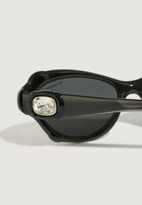Gafas de sol negras con un armazón brillante, que presentan un destacado acento de strass claro en la patilla. Lentes ahumadas grises con forma curvada.