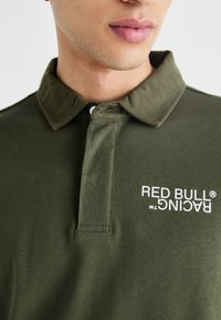 Olivno zelena polos majica izdelana iz gladkega bombažnega blaga, s ovratnikom na gumb in logotipom "RED BULL RACING" v beli barvi na prsih.