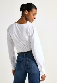 Camicia bianca a maniche lunghe con cuciture gather sulle spalle, abbinata a jeans blu aderenti con tasche posteriori visibili e dettagli in metallo dorato sui bottoni.