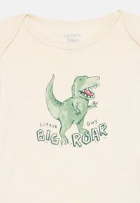 Mameluco de color crema con un dibujo de un dinosaurio verde y el texto "pequeño chico GRAN ROAR" en una fuente juguetona, confeccionado en suave material de algodón.