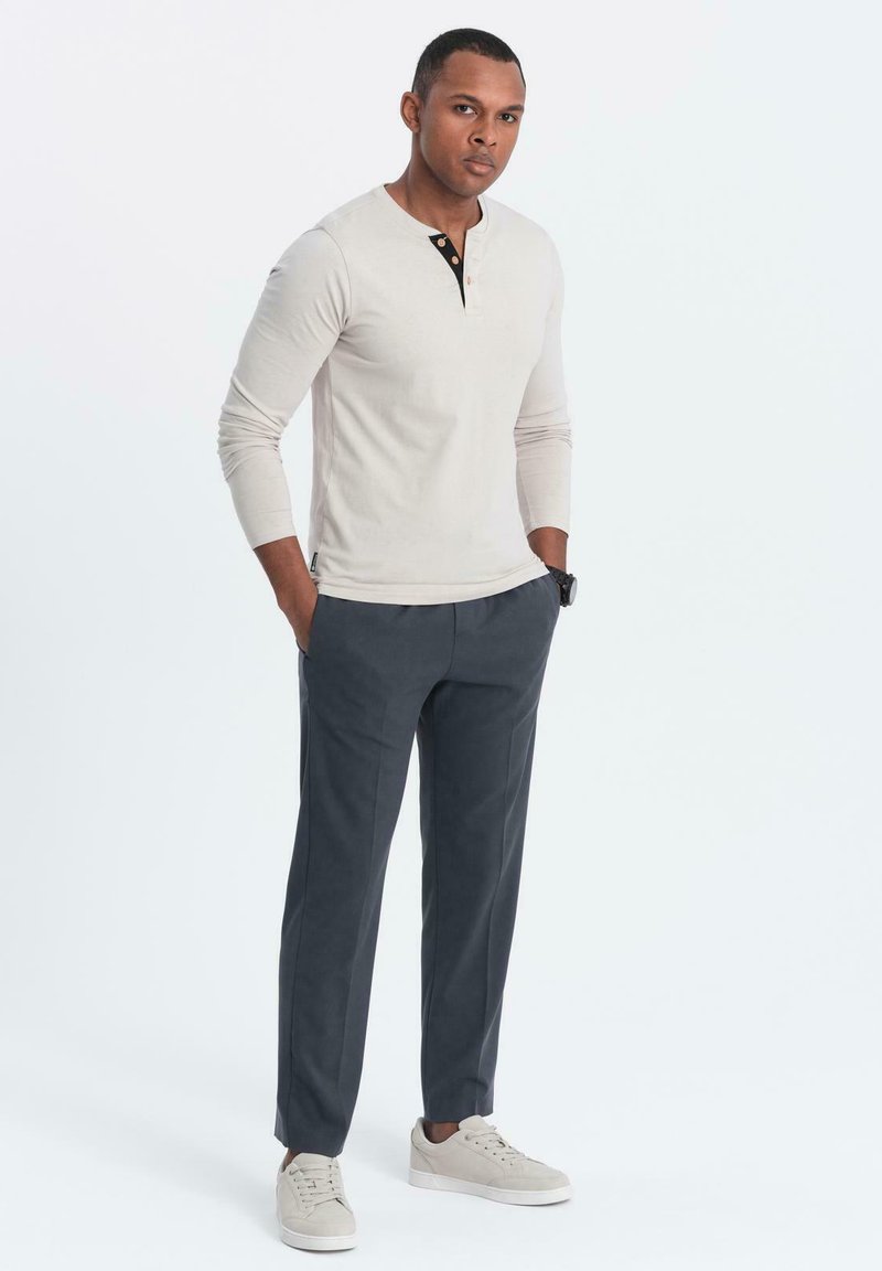 Maglietta henley a maniche lunghe beige chiaro con dettagli neri e bottoni, abbinata a pantaloni grigi scuri e sneakers grigie chiare. Sfondo neutro.