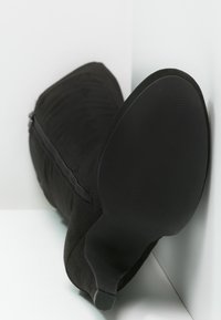 Chaussure à talons hauts en suede noir avec une texture lisse, un bout pointu et une fermeture éclair sur le côté. La semelle est en caoutchouc avec un motif en vagues.