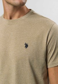 Beež lühikeste varrukatega T-särk, mis on valmistatud tekstuurse kangast, küljes väike tume logo vasakul rinnal, ümar kaelus ja vaba lõige.