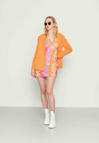 Blazer laranja sobre um vestido estampado em rosa e laranja, combinado com botas brancas até ao tornozelo e óculos de sol redondos pretos. Tecido suave, ajuste tailored.