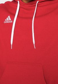 Punainen huppari, joka on valmistettu puuvillasekoitteesta, ja jossa on etutasku, valkoiset kiristysnyörit sekä pieni valkoinen Adidas-logo rinnassa.