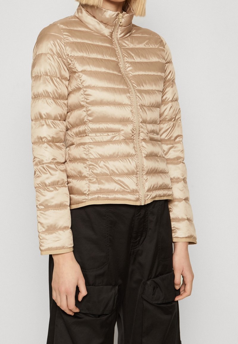 Beige Daunenjacke mit glänzender, gesteppten Oberfläche, hochstehendem Kragen, vorne mit Reißverschluss und zwei seitlichen Taschen. Getragen mit schwarzen Hosen.