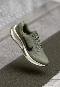 Αθλητικό παπούτσι τρεξίματος Nike σε ελαιί πράσινο με υφασμάτινη επιφάνεια με υφή, μαύρο λογότυπο swoosh και λευκή ενδιάμεση σόλα· διαθέτει περίπλοκο σχέδιο κορδονιών και στρογγυλεμένη μύτη.