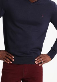 Pull col en V bleu marine en tissu tricoté, avec un petit logo brodé sur la poitrine, associé à un pantalon ajusté bordeaux.