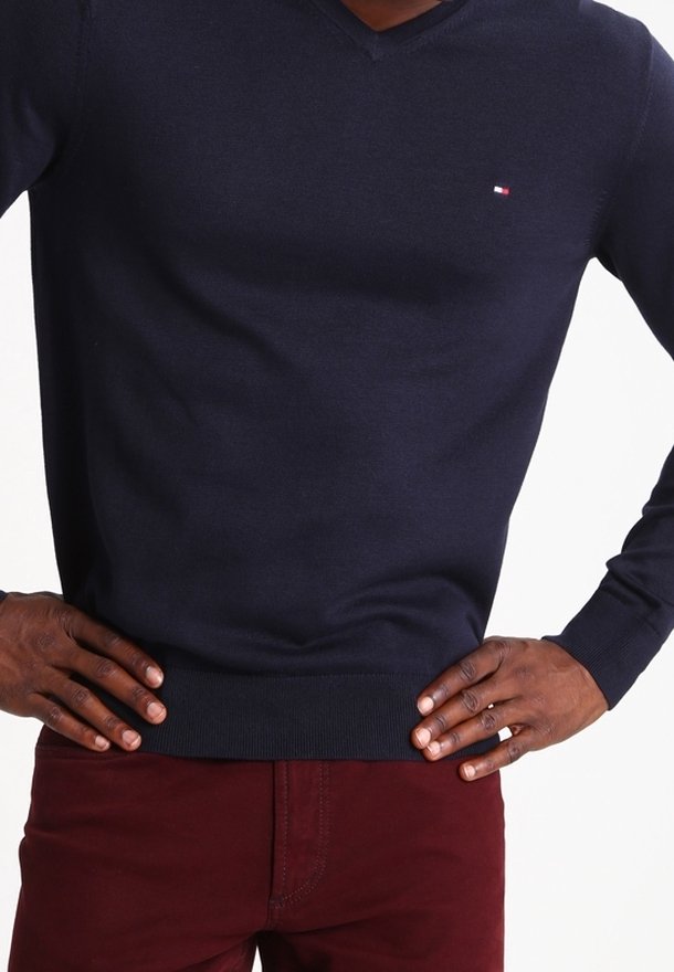 Pull col en V bleu marine en tissu tricoté, avec un petit logo brodé sur la poitrine, associé à un pantalon ajusté bordeaux.